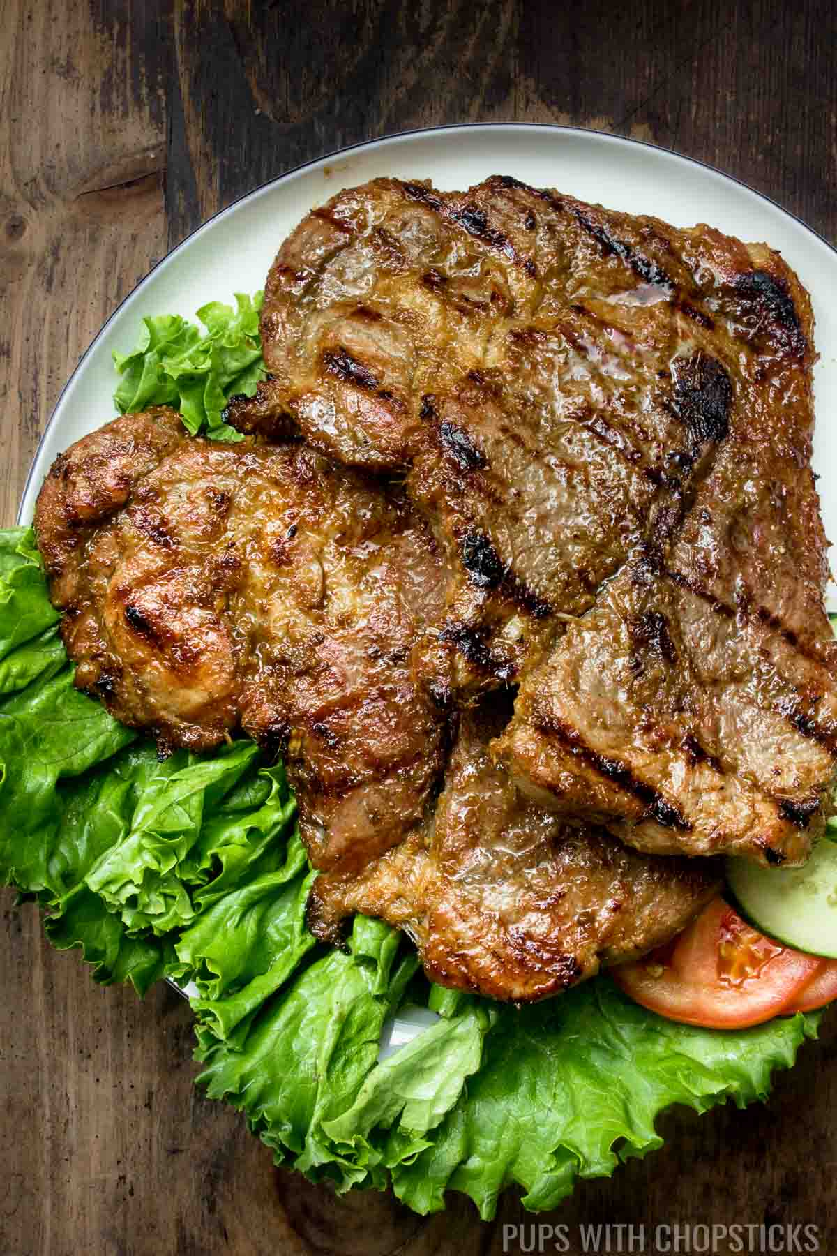 Two large pieces of grilled lemongrass pork chops on a white serving plate with tomatoes and cucumbers.