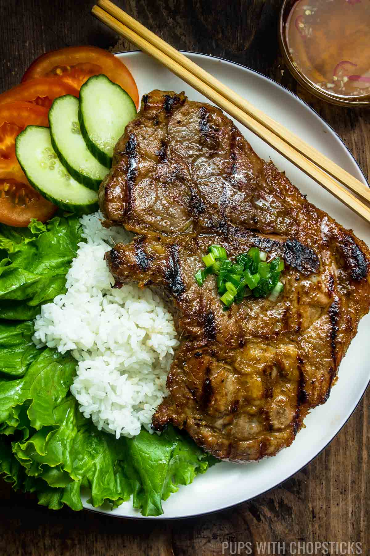 Vietnamese lemongrass pork chops served on a large white plate with rice, tomatoes and cucumber with a side of nuoc cham dipping sauce