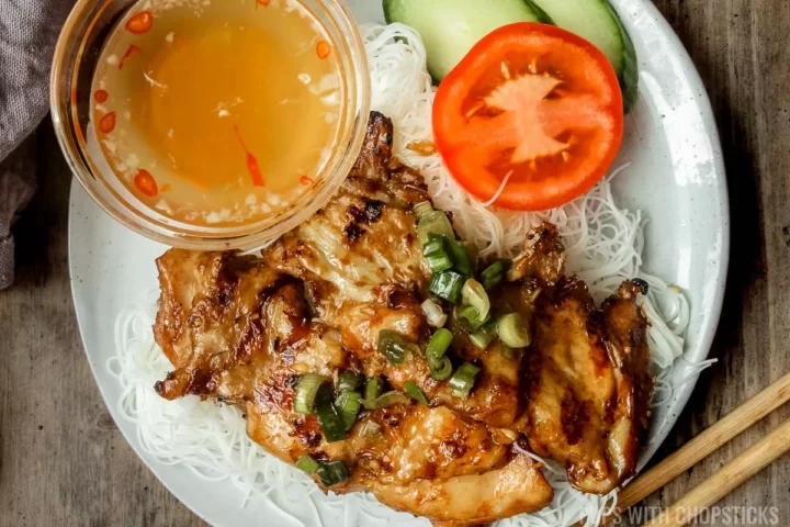 Lemongrass chicken served on vermicelli noodles, with a slice of cucumber and tomato and a side of nuoc cham.