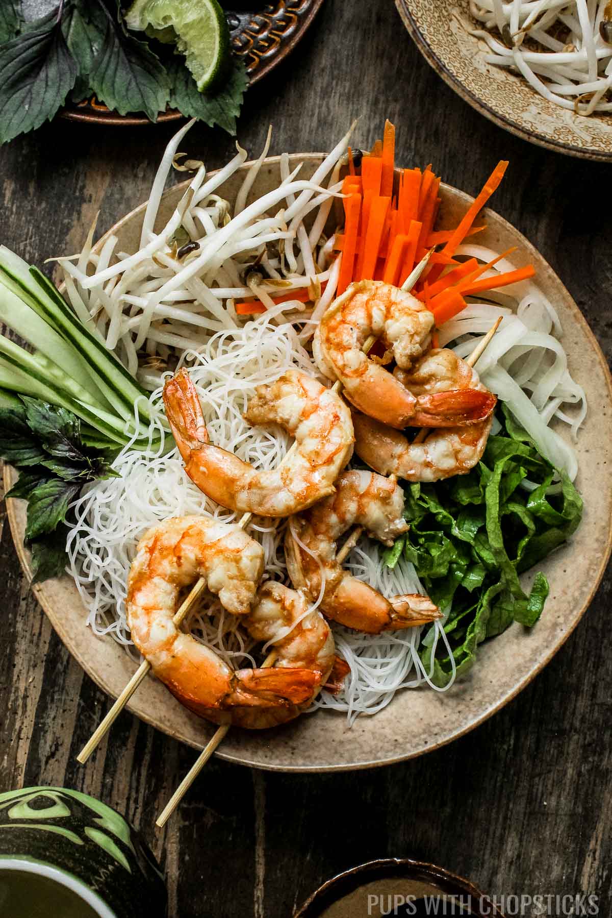 A bowl with vermicelli noodles, fresh vegetables and grilled shrimp on top.