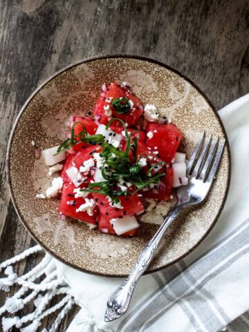 A small bowl with watermelon feta salad garnished with green onions and black sesame seeds and served with a fork