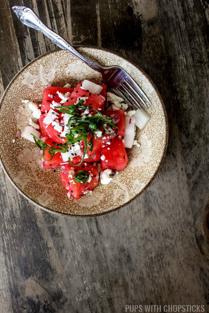 A small bowl with watermelon feta salad garnished with green onions and black sesame seeds and served with a fork