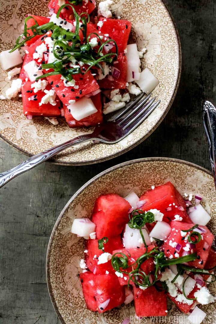 A close up of two small bowls with watermelon feta salad garnished with green onions and black sesame seeds and served with a fork