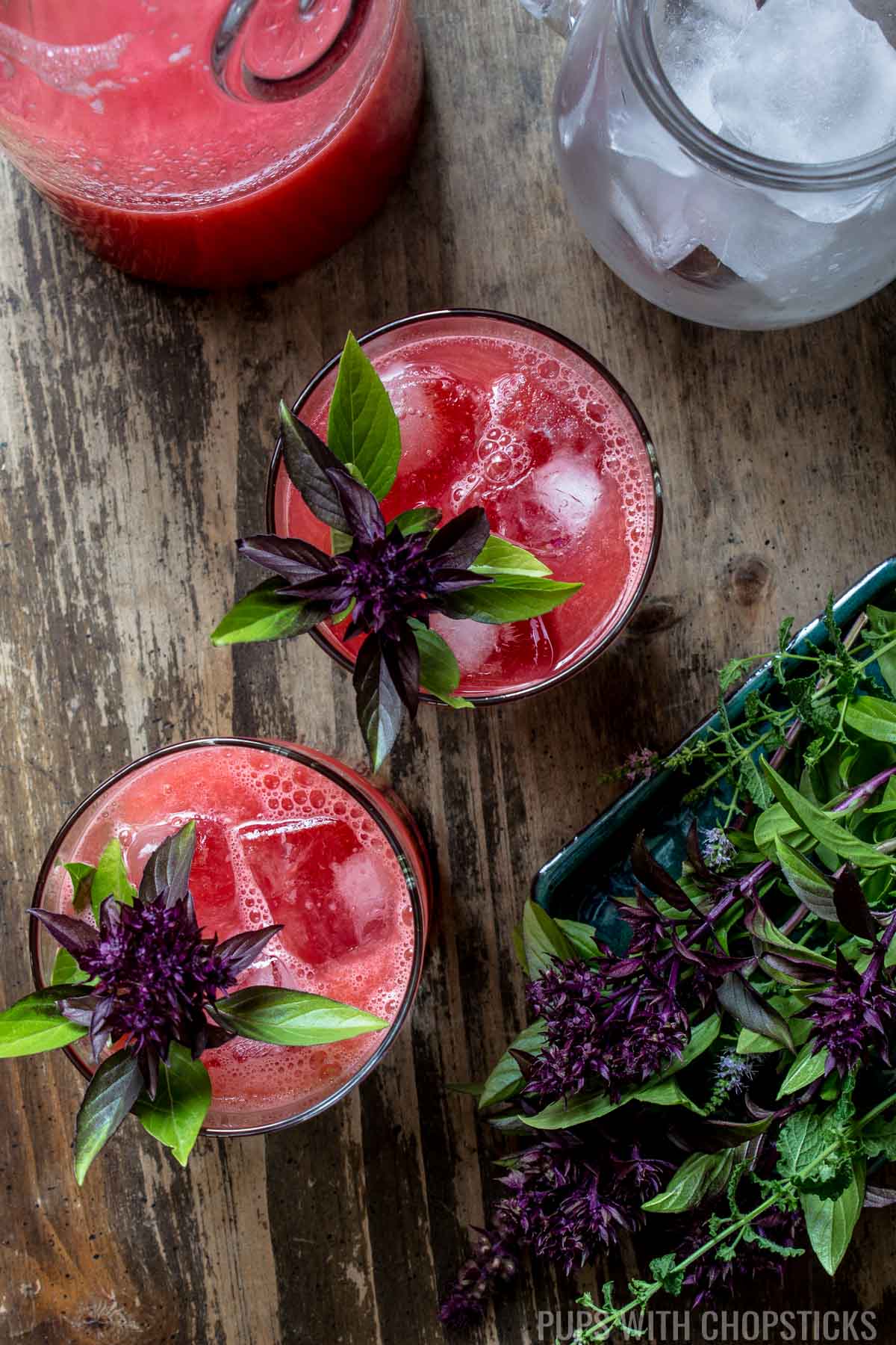 Two glasses of watermelon soju garnished with Thai basil and served with a jar of ice.