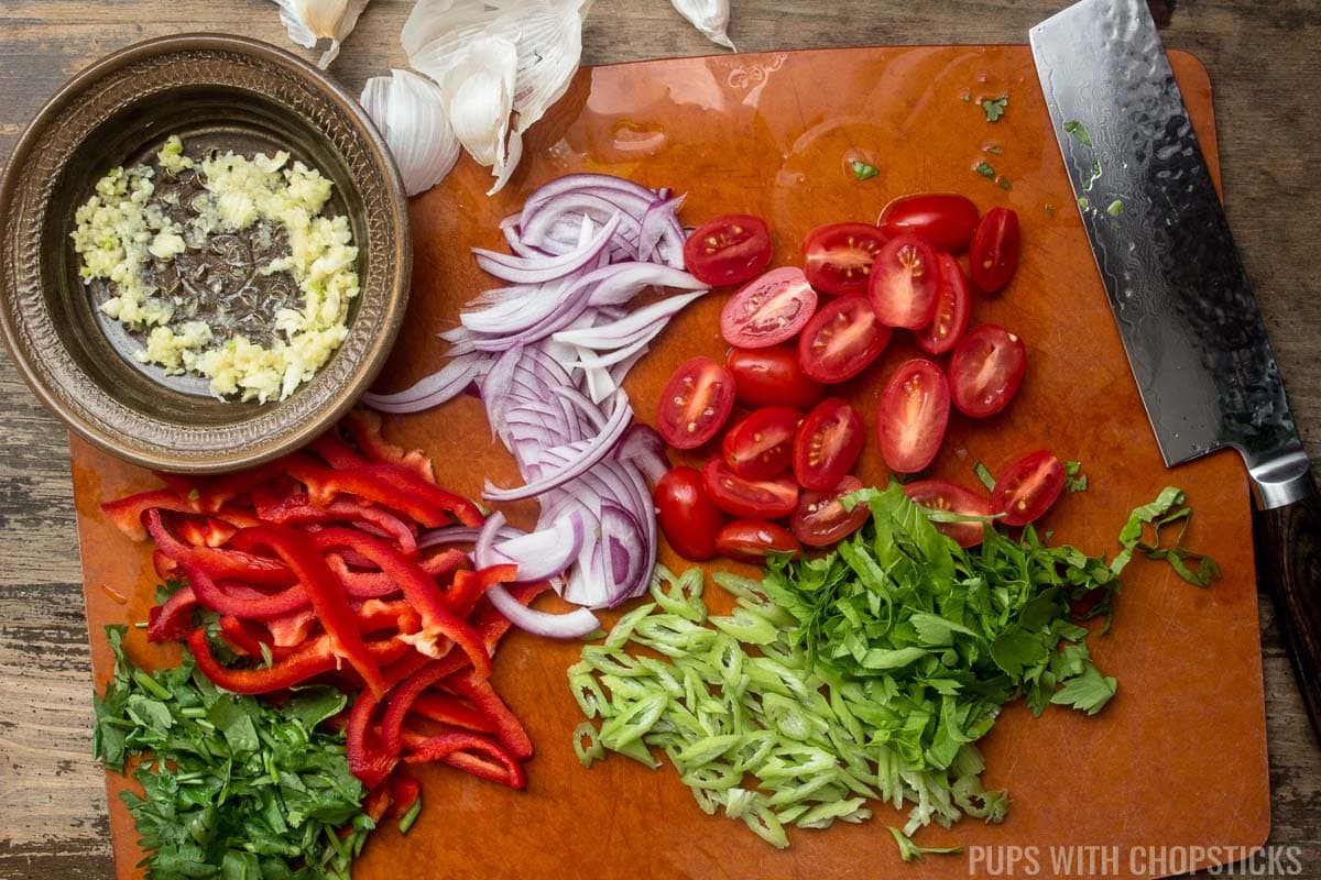 A cutting board with vegetables all chopped up and sliced (Chinese celery, baby tomatoes, red onions, red pepper, and parsley)